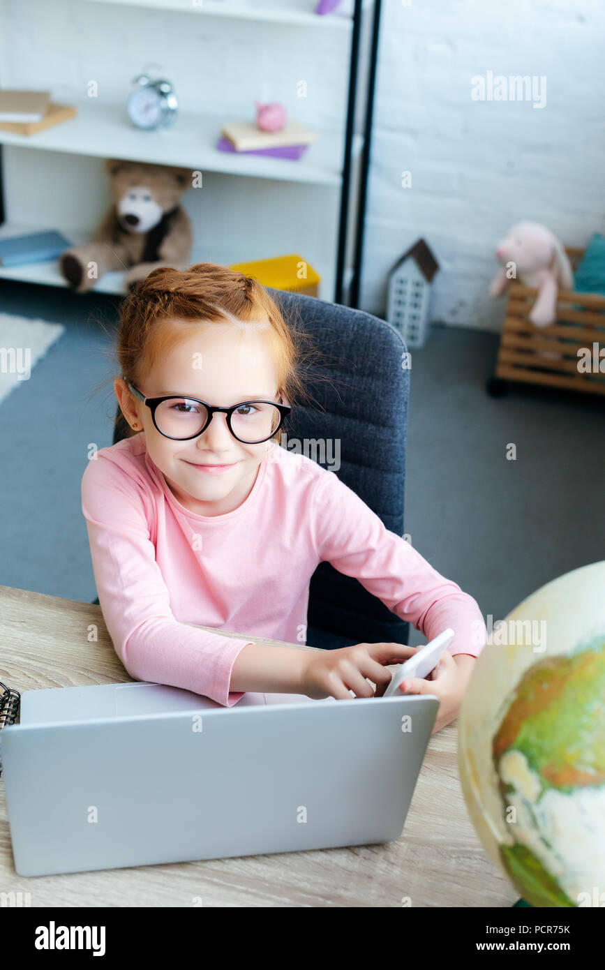 Hohe Betrachtungswinkel und niedlichen Rothaarigen Schulkind in Brillen in die Kamera lächeln, während Sie ihr Smartphone und Laptop Stockfoto