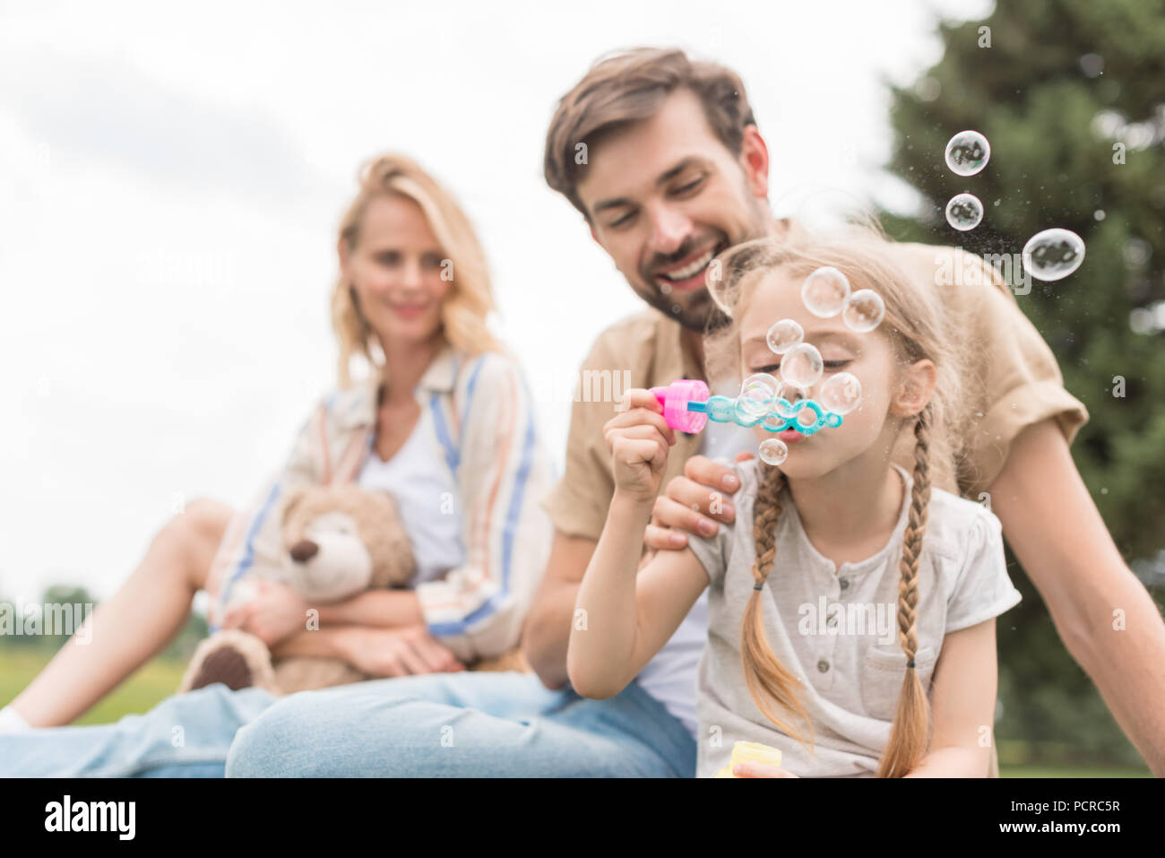 Mutter holding Teddybär und schauen sich Vater und Tochter bläst Seifenblasen in Park Stockfoto