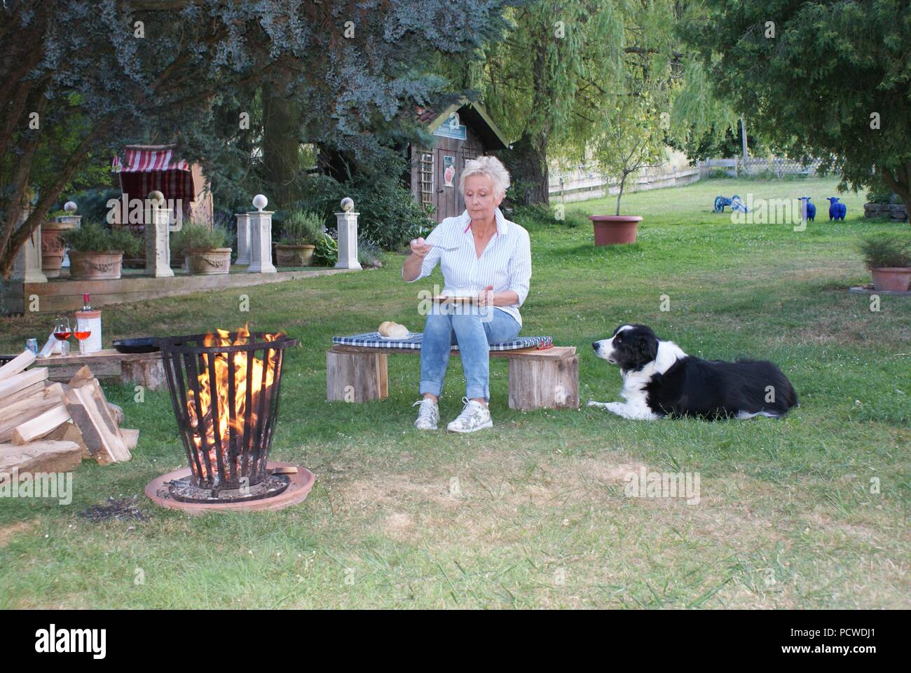 Die attraktive Frau isst Eier am Lagerfeuer gebraten, ihr Hund ist beobachten Stockfoto