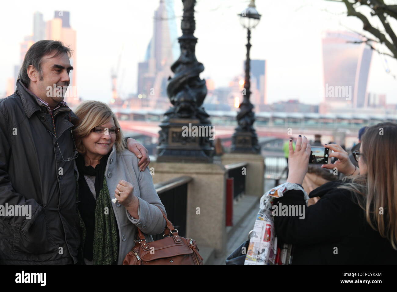 Mittleres Alter Paar posiert für ein Foto am Ufer der Themse mit Blick auf die Stadt im Hintergrund, als ihre Freundin nimmt Foto auf dem Smartphone. Stockfoto