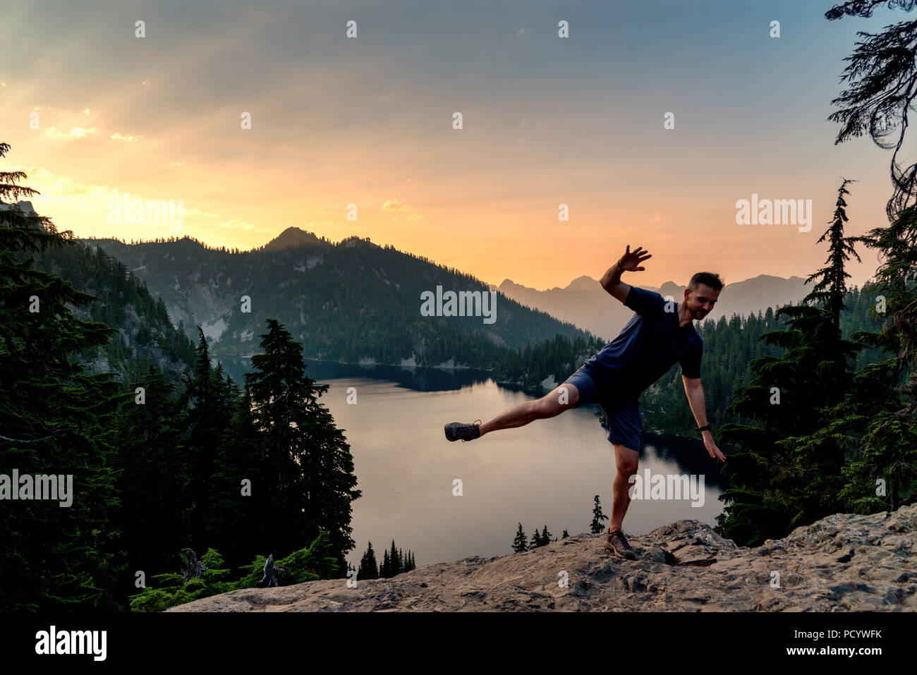 Humorvoll Mann verliert sein Gleichgewicht, während auf einer Klippe mit Blick auf den See bei Sonnenuntergang posing Stockfoto