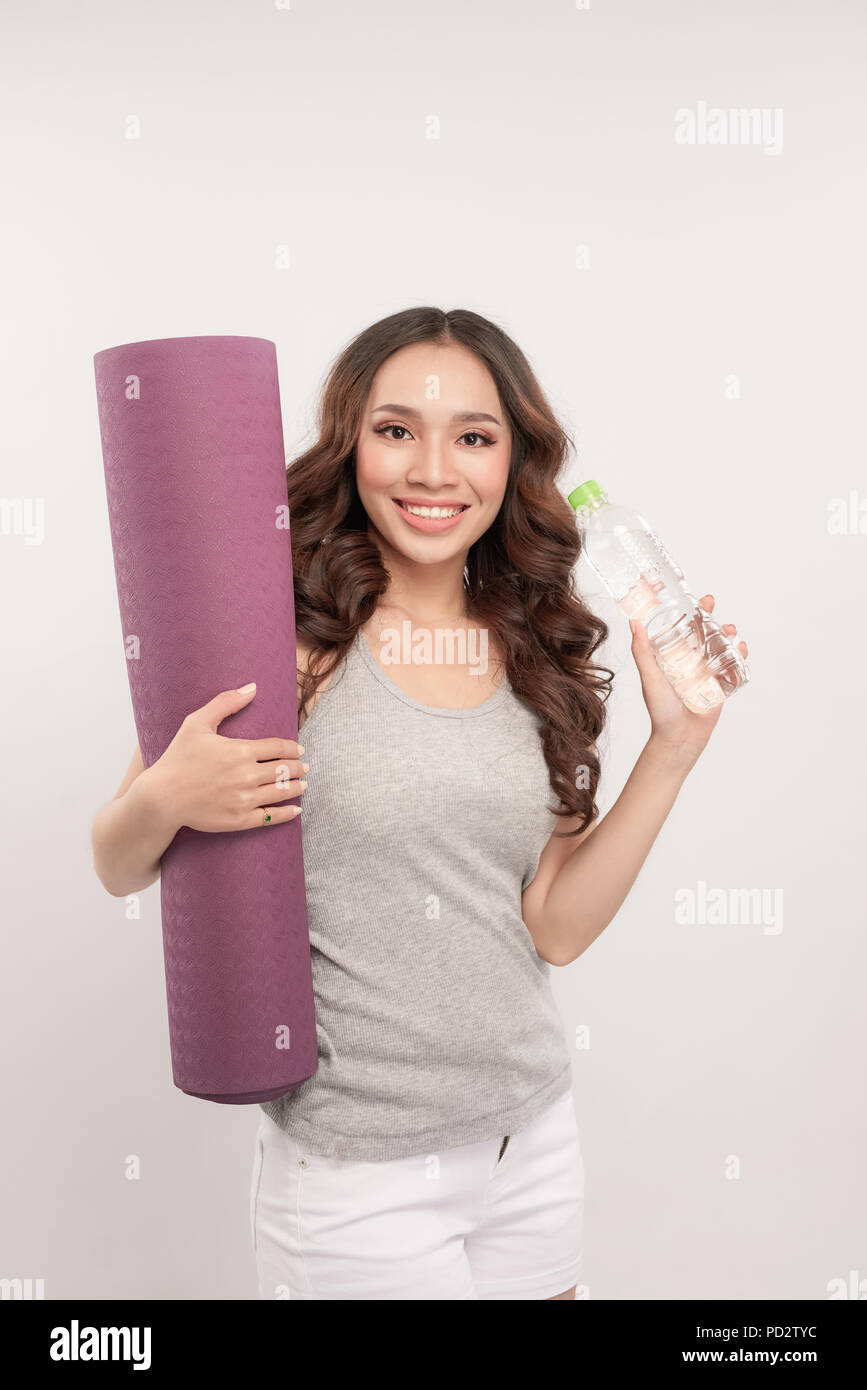 Schöne junge Frau in Sportbekleidung hält eine Yogamatte und eine Flasche Wasser, in die Kamera schauen und lächeln, stehend auf einem grauen Hintergrund Stockfoto