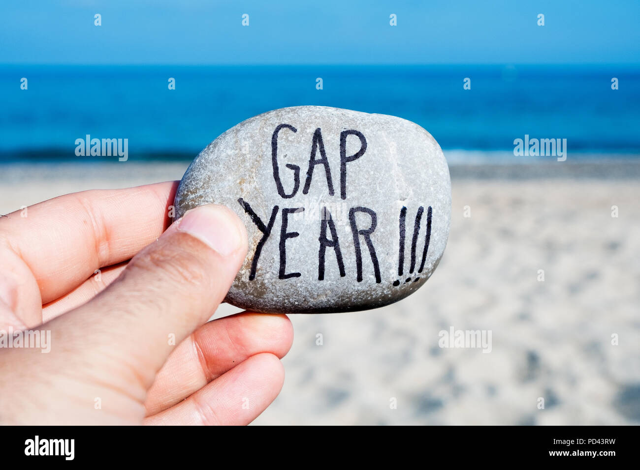 Nahaufnahme der Hand eines jungen kaukasischen Mann am Strand, vor dem Ozean, mit einem Stein mit dem Text gap year geschrieben Stockfoto