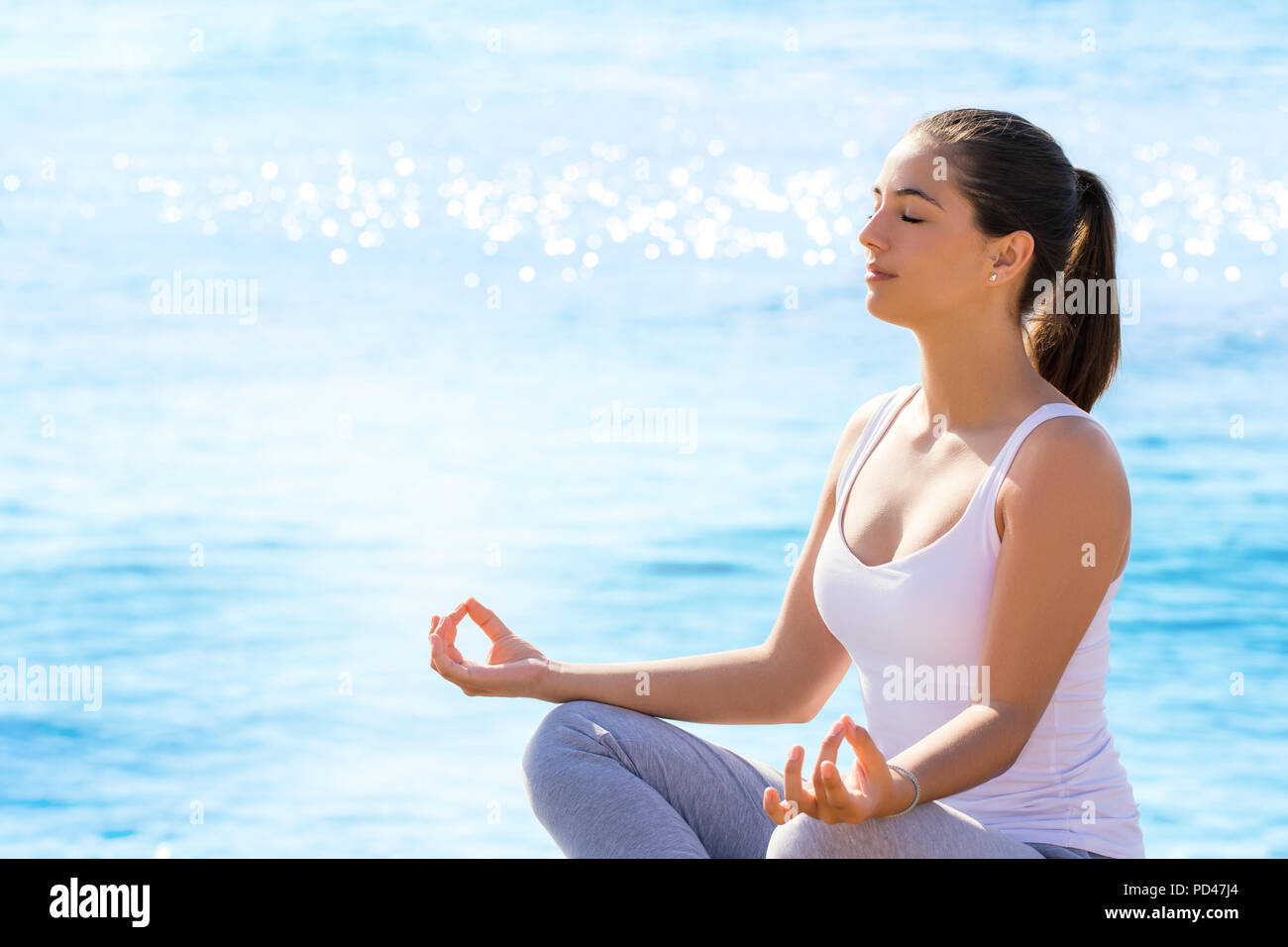 Nahaufnahme, Porträt der jungen Frau, meditieren, Yoga Position im Freien. Mädchen in Freizeitkleidung sitzt gegen den blauen Meer Hintergrund mit Sun auf reflektierenden Stockfoto