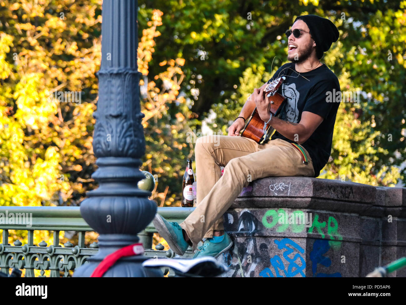 Straßenmusiker spielt Gitarre auf einer Brücke in Kreuzberg, Berlin, umgeben von Graffiti und Herbstfarben, die eine entspannte urbane Atmosphäre schaffen. Stockfoto
