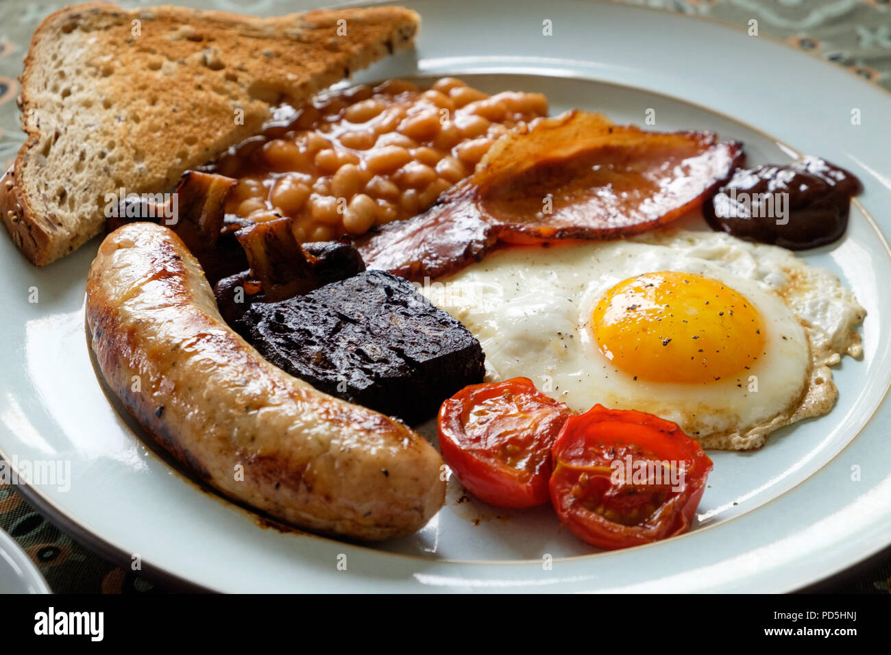 Ein hausgemachtes englisches Frühstück mit Ei, Speck, Cumberland Würstchen, Blutwurst, gebackene Bohnen, Tomaten, Pilze, Toast und eine Tasse Kaffee. Stockfoto