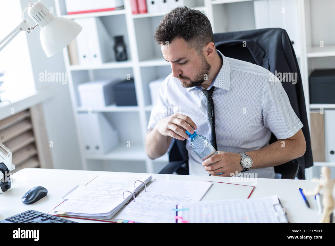Ein Mann sitzt an einem Tisch im Büro, Arbeiten mit Dokumenten und öffnen eine Flasche Wasser. Stockfoto