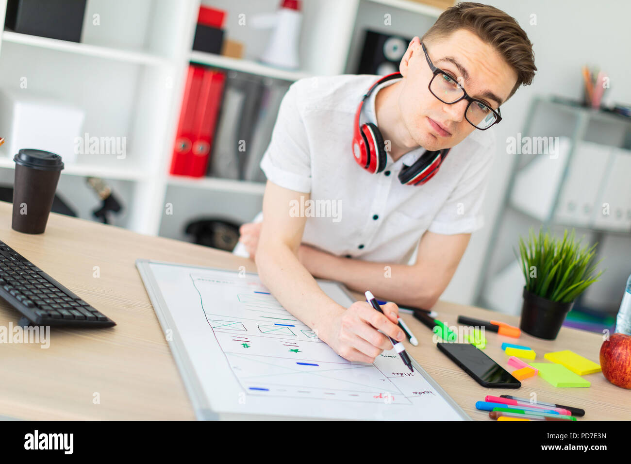 Ein junger Mann in Gläsern steht in der Nähe ein Computer Schreibtisch. Ein junger Mann zeichnet eine Markierung auf einem magnetischen Brett. Auf dem Hals, die Kopfhörer der Kerl hängen. Stockfoto