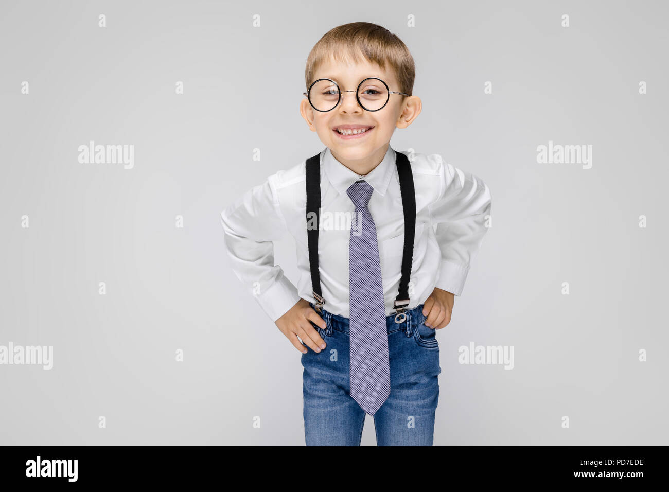 Eine charmante junge in ein weißes Hemd, Hosenträger, krawatte und leichte Jeans steht auf einem grauen Hintergrund. Der Junge mit Brille lächeln Stockfoto