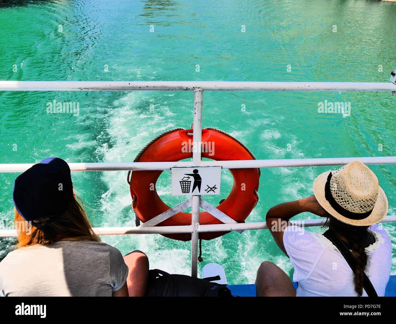 Hoher Kontrast Rückansicht 2 Junge weibliche Reisende in sunhats sitzen auf See Komani Fähre, Albanien, bewundern, ein roter Rettungsring & keine Einstreu unterzeichnen. Stockfoto