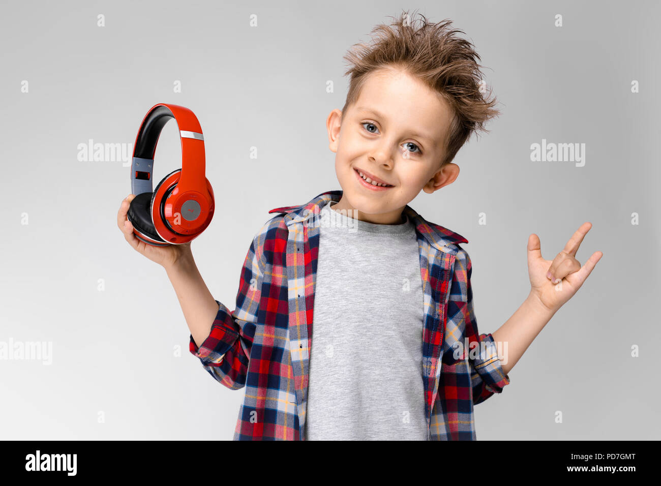 Ein schöner Junge in einem Plaid Shirt, grau Shirt und Jeans steht auf einem grauen Hintergrund. Ein Junge in rot Kopfhörer. Der Junge nahm seinen Kopfhörer. Stockfoto