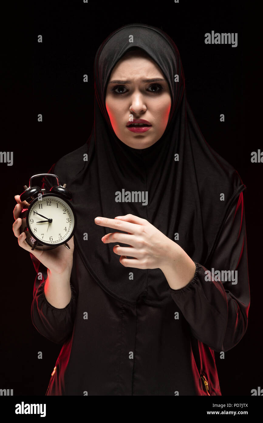 Porträt der schönen schweren Angst junge muslimische Frau mit schwarzen Hijab holding Wecker auf schwarzem Hintergrund Angst Stockfoto