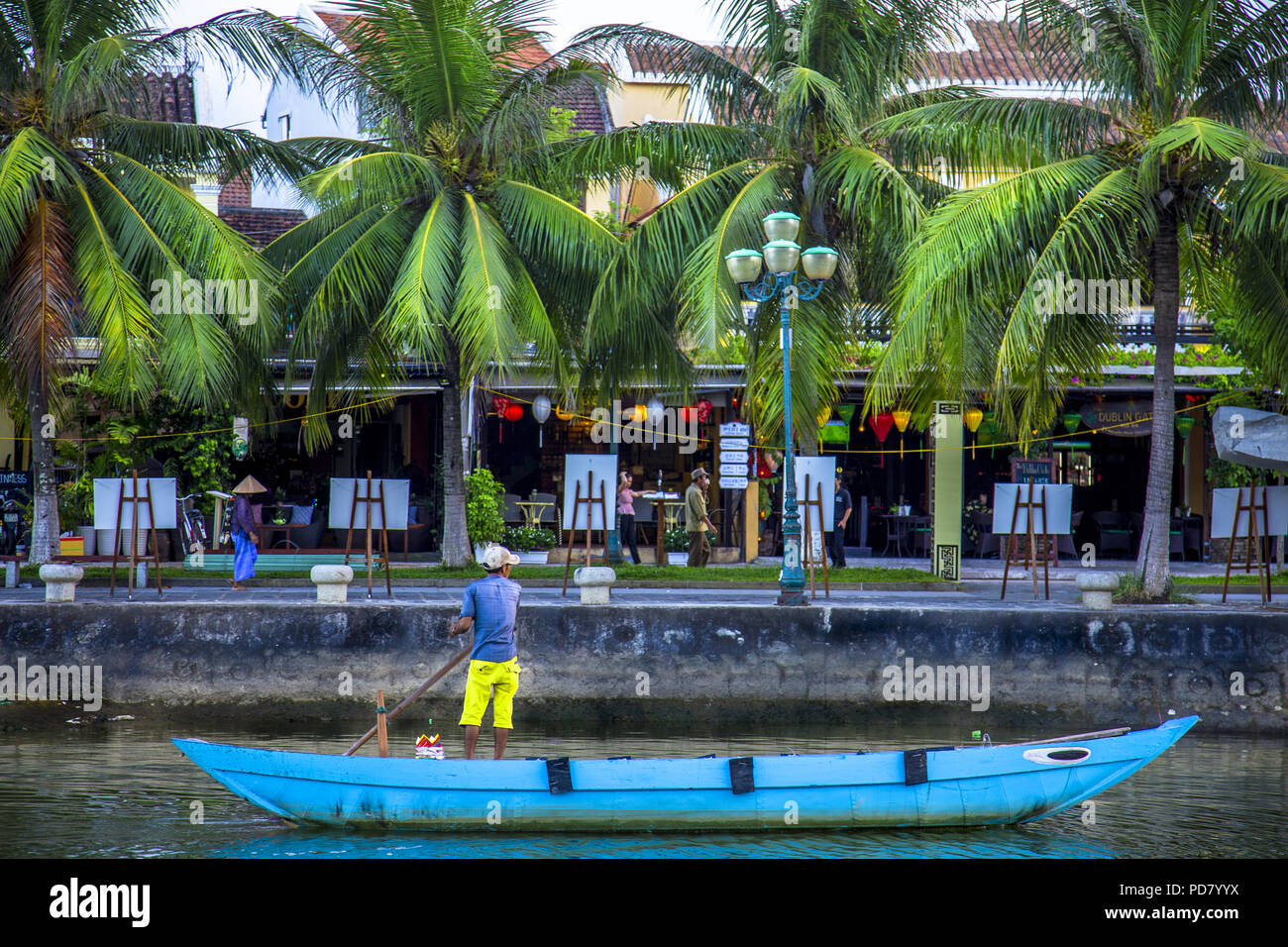 Ende Tag Farben mit blauen Boot und Mann mit einem Pol in sein Boot. Der Hintergrund hat lebhaftes Grün Palmen vor Hoi An Store Fronten. Freigabe Stockfoto