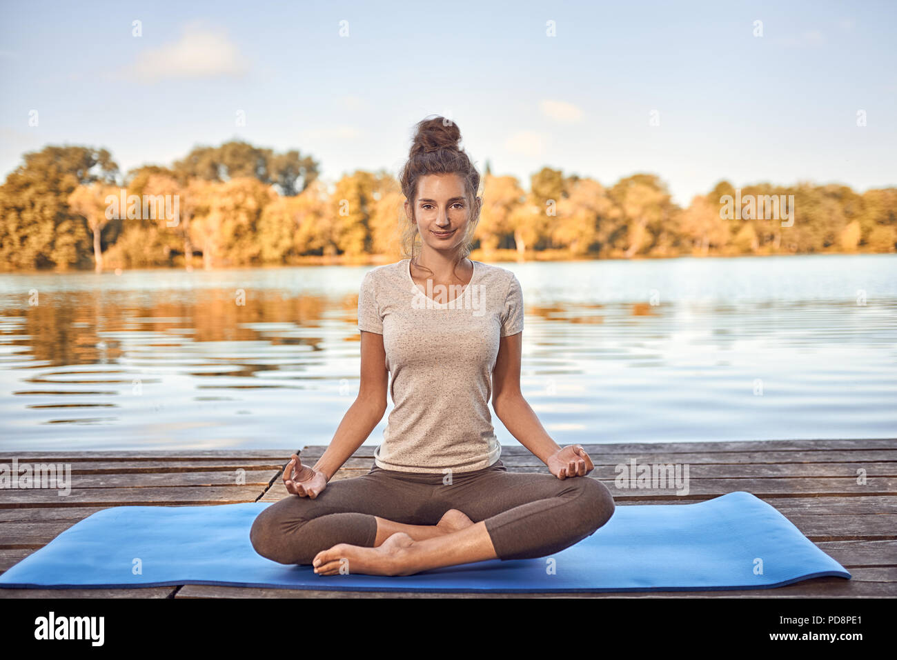 Lächelnde Frau sitzt auf einem Yoga Matte im Lotussitz meditierend auf einem holzdeck oder Steg mit Blick auf einen ruhigen See in einen gesunden Lebensstil conc Stockfoto