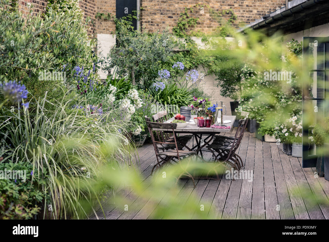 Alte französische Bistro Stühle um langen Holztisch auf Garten Dachterrasse mit Belag. Agapanthus und Jasmin blüht im Hintergrund Stockfoto