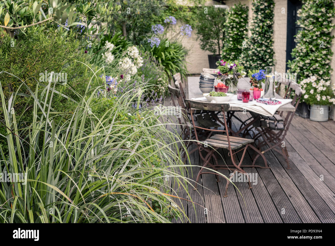 Alte französische Bistro Stühle um langen Holztisch auf Garten Dachterrasse mit Belag. Agapanthus und Jasmin blüht im Hintergrund Stockfoto