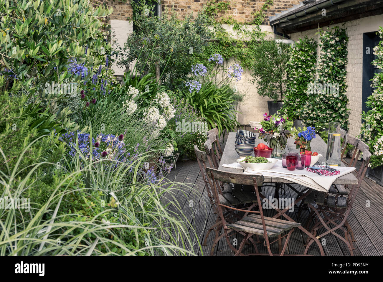 Alte französische Bistro Stühle um langen Holztisch auf Garten Dachterrasse mit Belag. Agapanthus und Jasmin blüht im Hintergrund Stockfoto