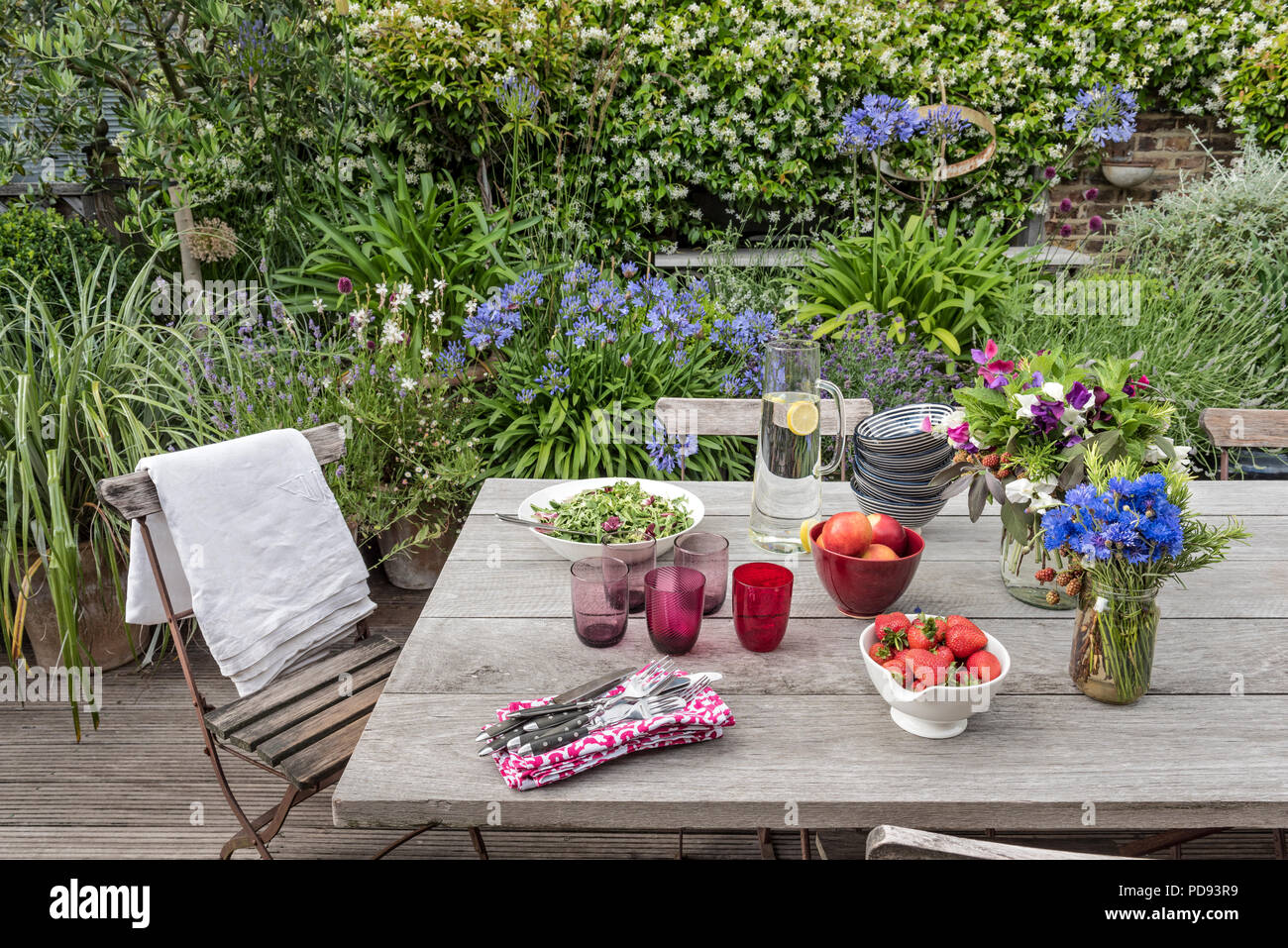 Rustikalen Holztisch auf Garten Dachterrasse für ein leichtes Mittagessen im Freien. Agapanthus und Jasmin im Hintergrund blühen Stockfoto