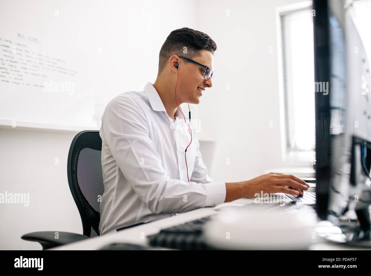 Lächelnd Software Entwickler sitzt in seinem Büro Schreibtisch arbeiten am Laptop das Tragen von Kopfhörern. Mann mit Brille arbeiten am Laptop im Büro Stockfoto