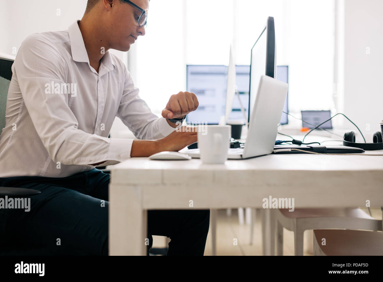 Software Entwickler sitzt in seinem Büro Schreibtisch, die auf Notebook- und Desktop-Computer, die gleichzeitig arbeiten. Mann Tragen einer Brille auf seine Armbanduhr suchen Stockfoto