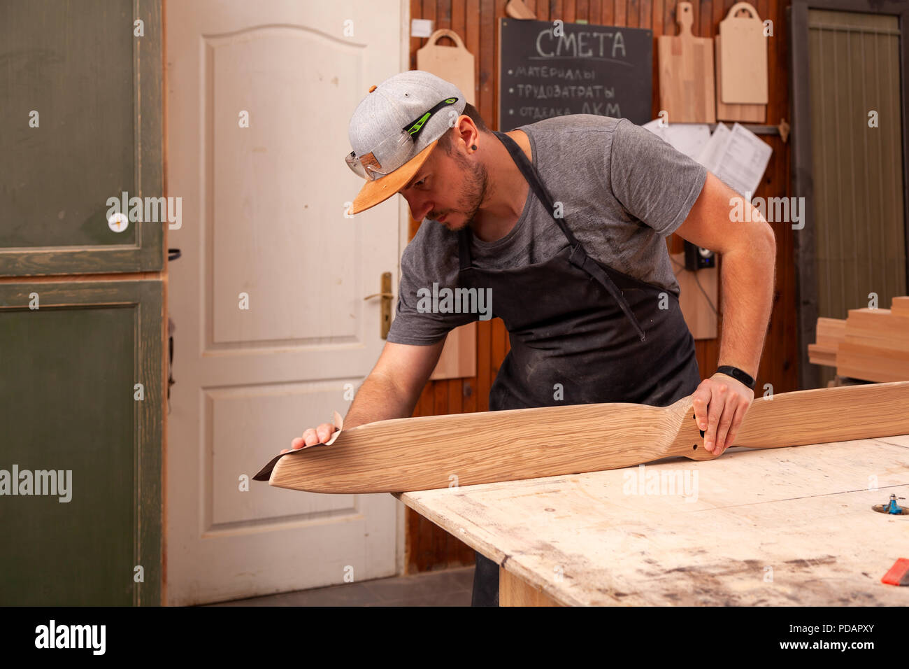 Ein Mann in einem Cap und Shirt poliert die Holzblock mit Sandpapier vor dem Lackieren in der Werkstatt, im Hintergrund, Werkzeuge und Bohren mach Stockfoto