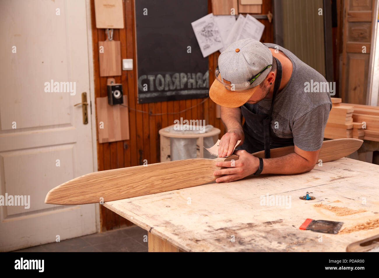 Ein Mann in einem Cap und Shirt poliert die Holzblock mit Sandpapier vor dem Lackieren in der Werkstatt, im Hintergrund, Werkzeuge und Bohren mach Stockfoto