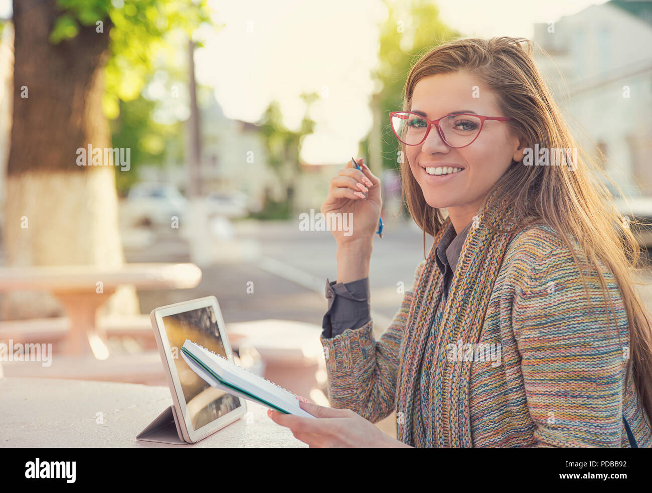 Hübsche stilvolle hipster Frau in Gläsern sitzen auf der Straße mit Tablet und Notepad Studien in und lächelnd an Kamera Stockfoto