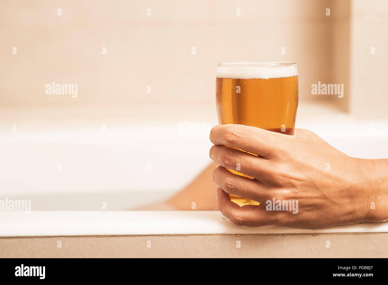 Nahaufnahme eines jungen kaukasischen Mann trinkt ein erfrischendes Bier, während sich bei einem Bad in der Badewanne von einem beigefarbenen Badezimmer Stockfoto