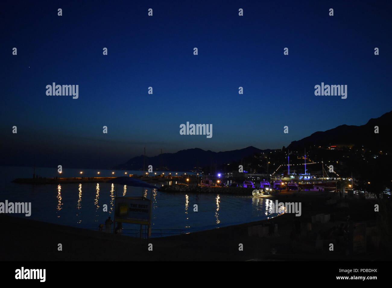 Nacht von Baska Voda Hafen, Strand, Boote und Geschäfte mit Elektrischen blauen Himmel, die Berge im Hintergrund und Lichtreflexe im Ozean Stockfoto