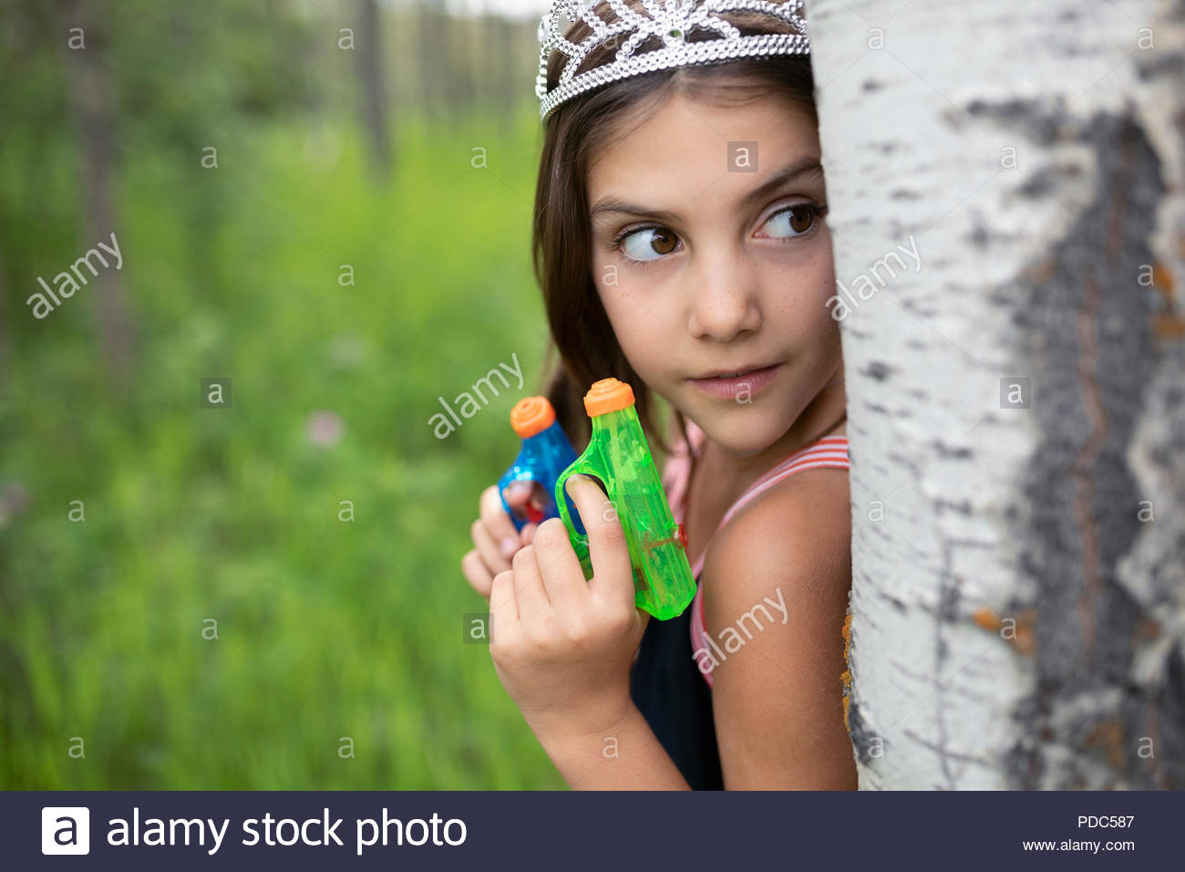 Mädchen in der Tiara mit sprizen Gewehren versteckt sich hinter Baum Stockfoto