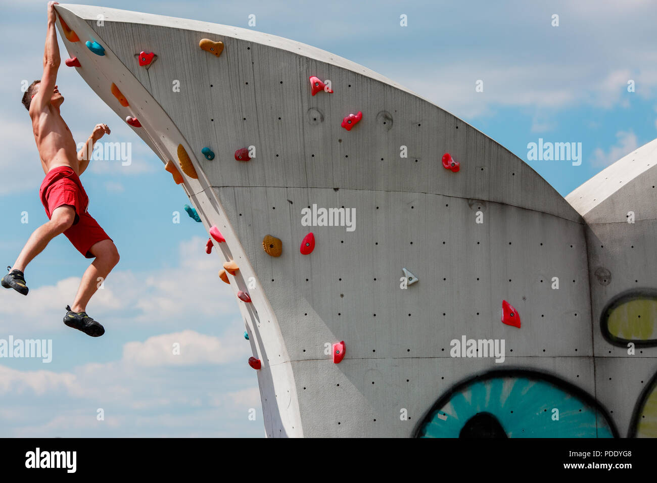 Foto von starken Sport brunet in roten Hosen hängen an der Wand zum Klettern gegen den blauen Himmel mit Wolken Stockfoto