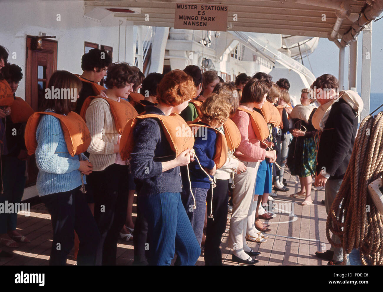 1960, historische Gruppe junger Leute stehen draussen auf dem Deck eines Schiffes in orange Schwimmwesten bereit, sich in einer Sicherheit Bohrer zu nehmen. Stockfoto