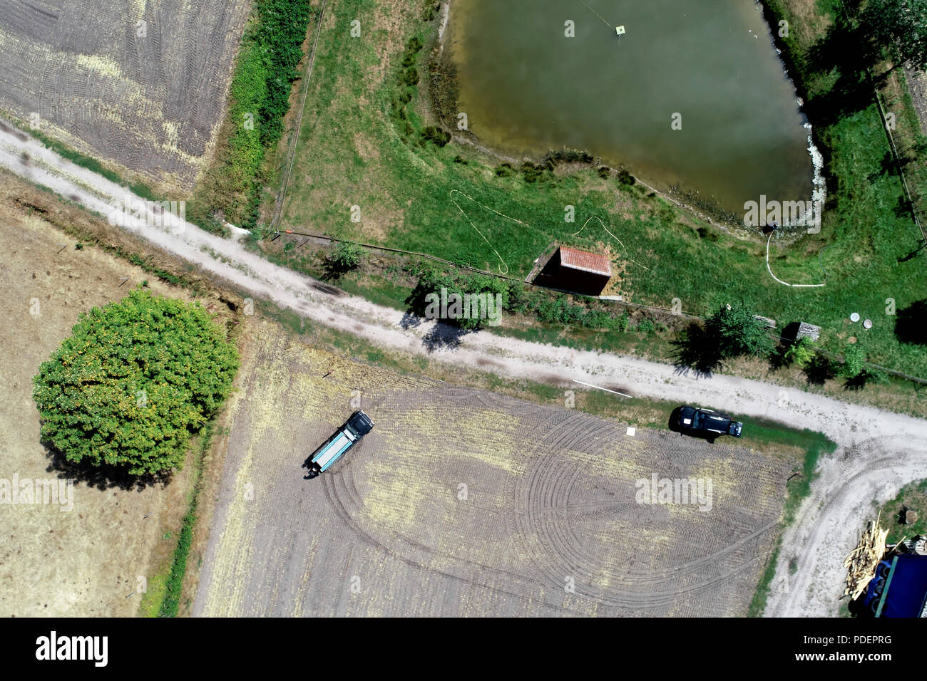 Vertikale Luftaufnahme über einen Weg neben einem Abgeernteten Feld und eine Wiese mit einem kleinen Stall vor einem Teich zum Angeln. Stockfoto