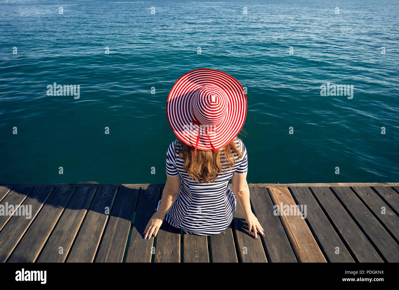 Frau mit Hut und gestreiften Kleid sitzt auf dem Pier und dem türkisfarbenen Meer. Sommer und Meer Konzept. Stockfoto