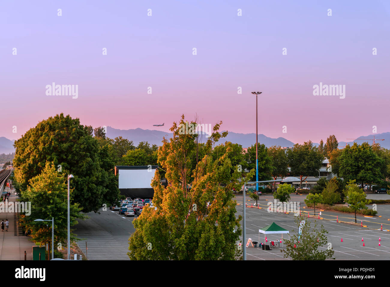 Parkplatz mit Sommerkino, grüne Bäume, Berge, Fliegen, Flugzeug, Autos auf Sommerabend in der modernen Stadt Stockfoto