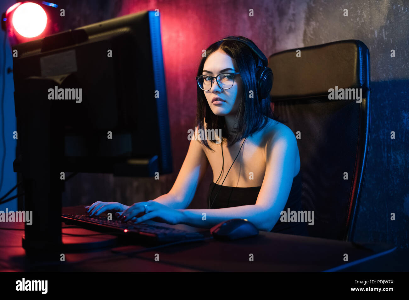 Schöne junge Mädchen mit Brille und Gaming Headset spielt online Spiel auf Gaming-PC im dunklen Bereich. Streaming online Spiele auf Internet Stockfoto