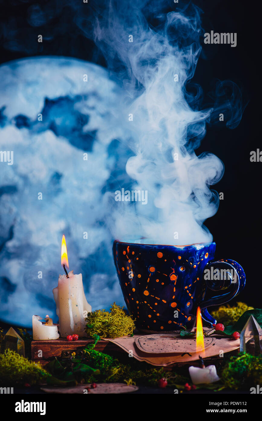Blau Keramiktasse mit Sternbilder und Sterne in einer dunklen noch leben. Magier Arbeitsplatz mit brennenden Kerzen, Rauch, Kristalle, Moos und trank Flaschen Stockfoto