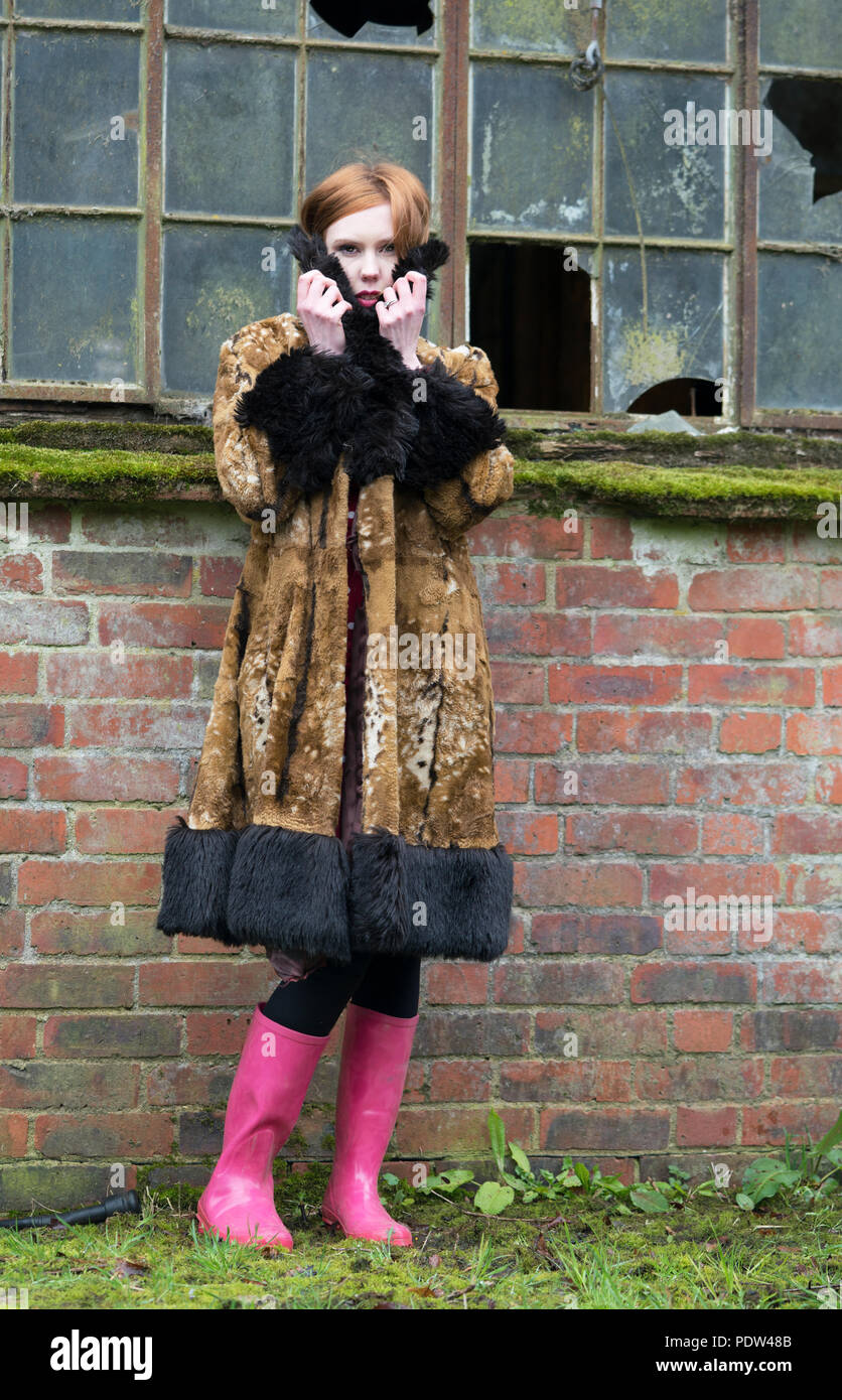 Rothaarige Mädchen in Braun vintage faux Fell mit schwarzen Zierleisten und rosa Stiefel sieht das Einfrieren vor einem verlassenen Bauernhof mit Glasscherben. Stockfoto