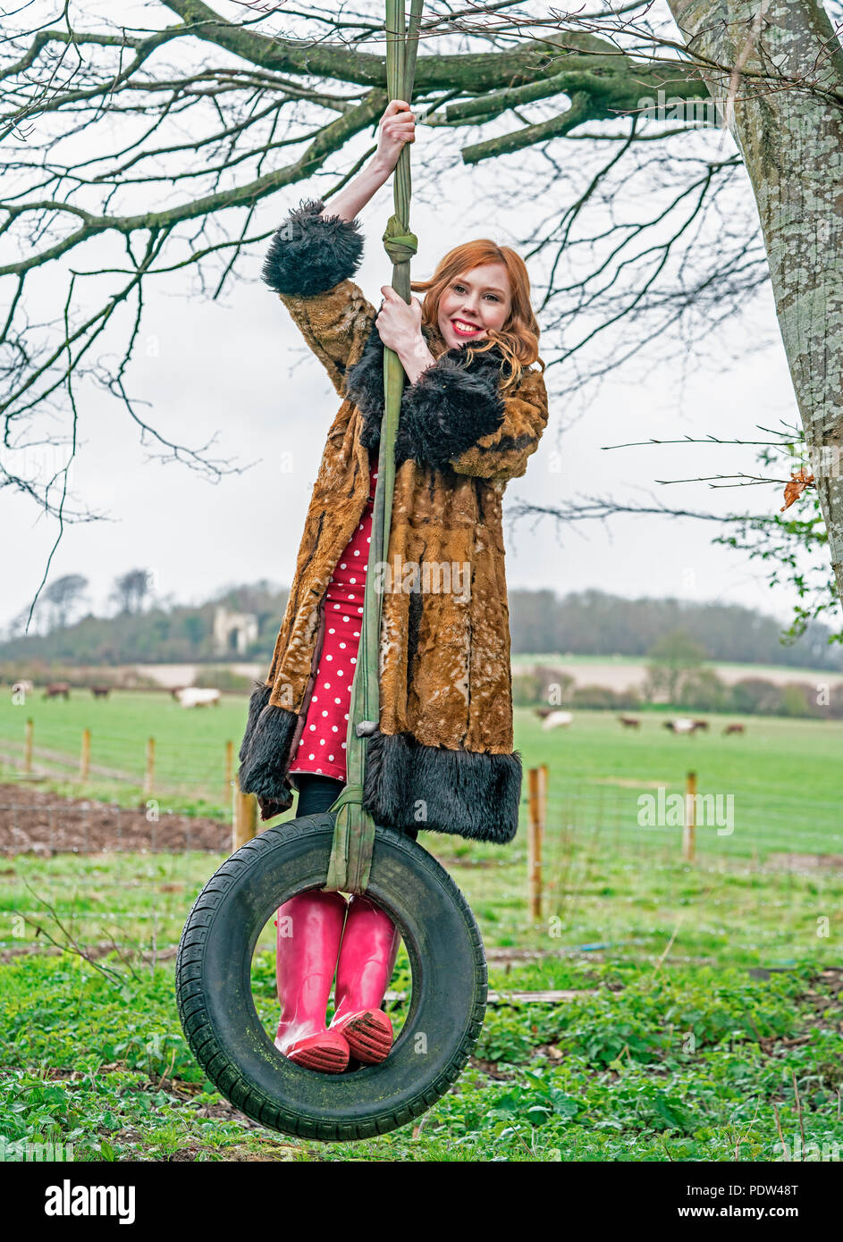 Rothaarige Dame in vintage Fellimitat Mantel, rot gepunkteten Kleid und rosa Gummistiefel schwingt von einem Baum auf einem Gummireifen auf der Farm. Stockfoto
