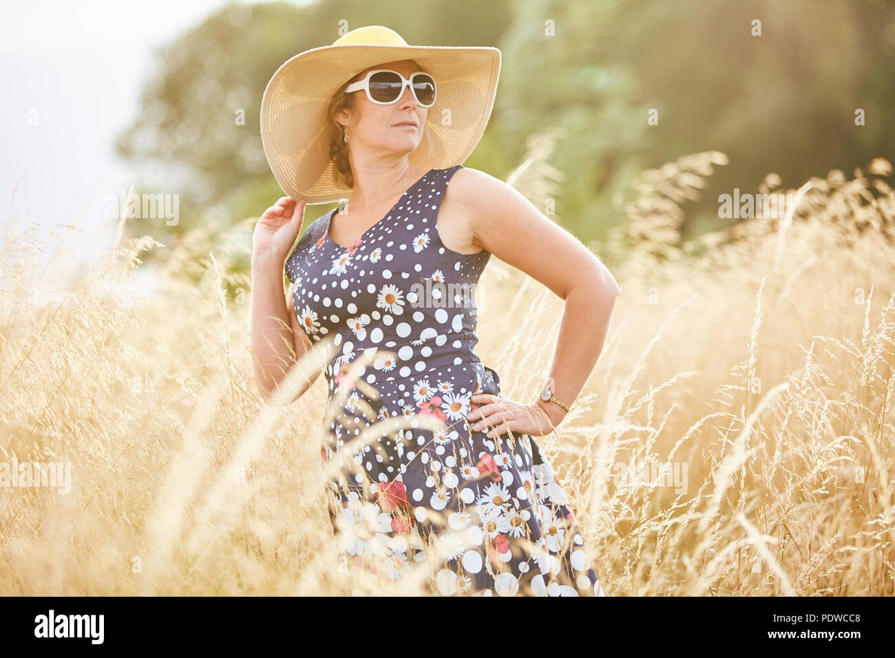 Frau mittleren Alters mit einem breitrandigen Hut, stehend in langen whispy golden Grass während der Suche mit Sonnenbrille auf der rechten Seite auf einem hellen Tag Stockfoto