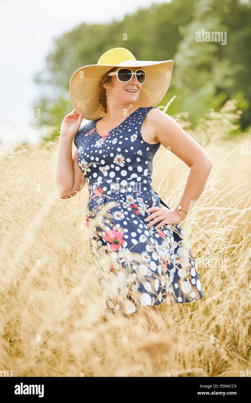 Frau mittleren Alters mit einem breitrandigen Hut, stehend in langen whispy golden Grass während der Suche mit Sonnenbrille auf der rechten Seite auf einem hellen Tag Stockfoto