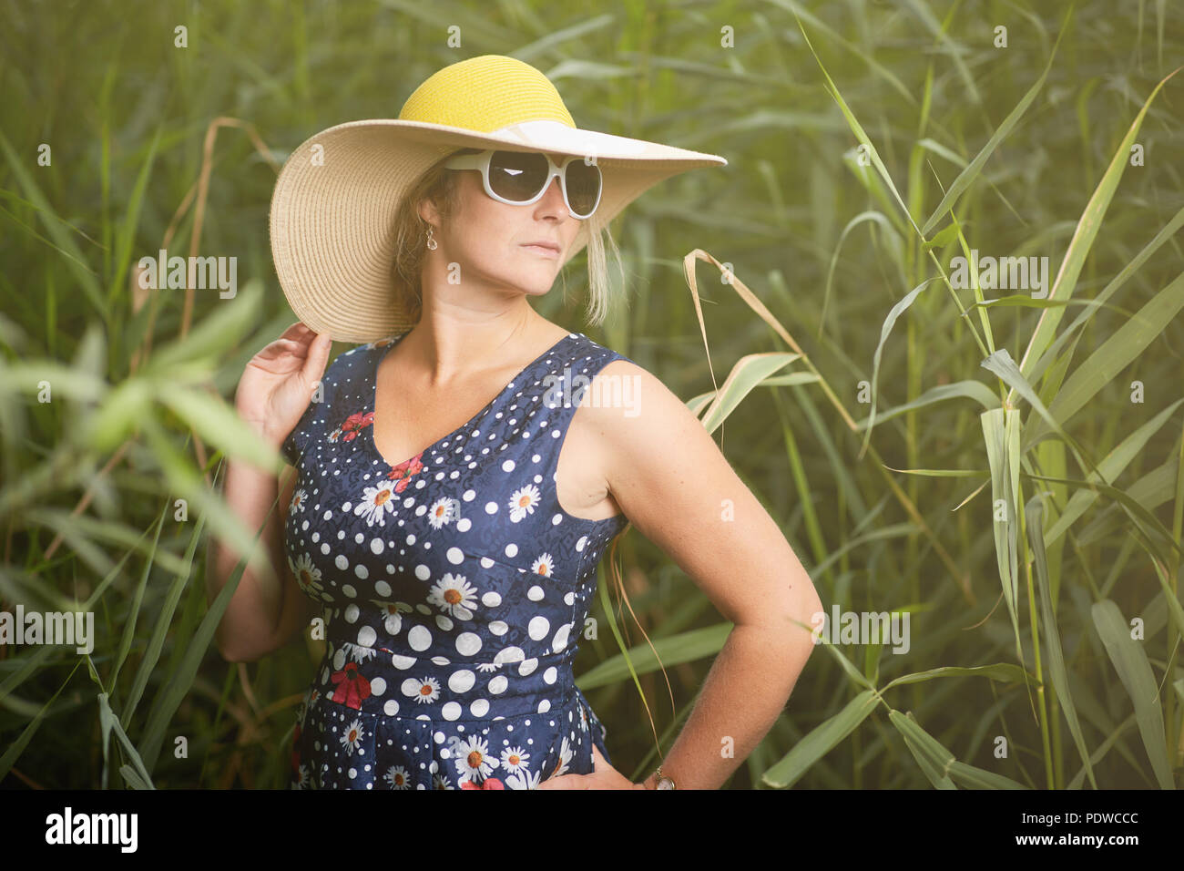 Frau mittleren Alters tragen weiße Sonnenbrille mit breiter Krempe stehend hat unter langen grünen Gras, auf der Suche nach rechts Stockfoto