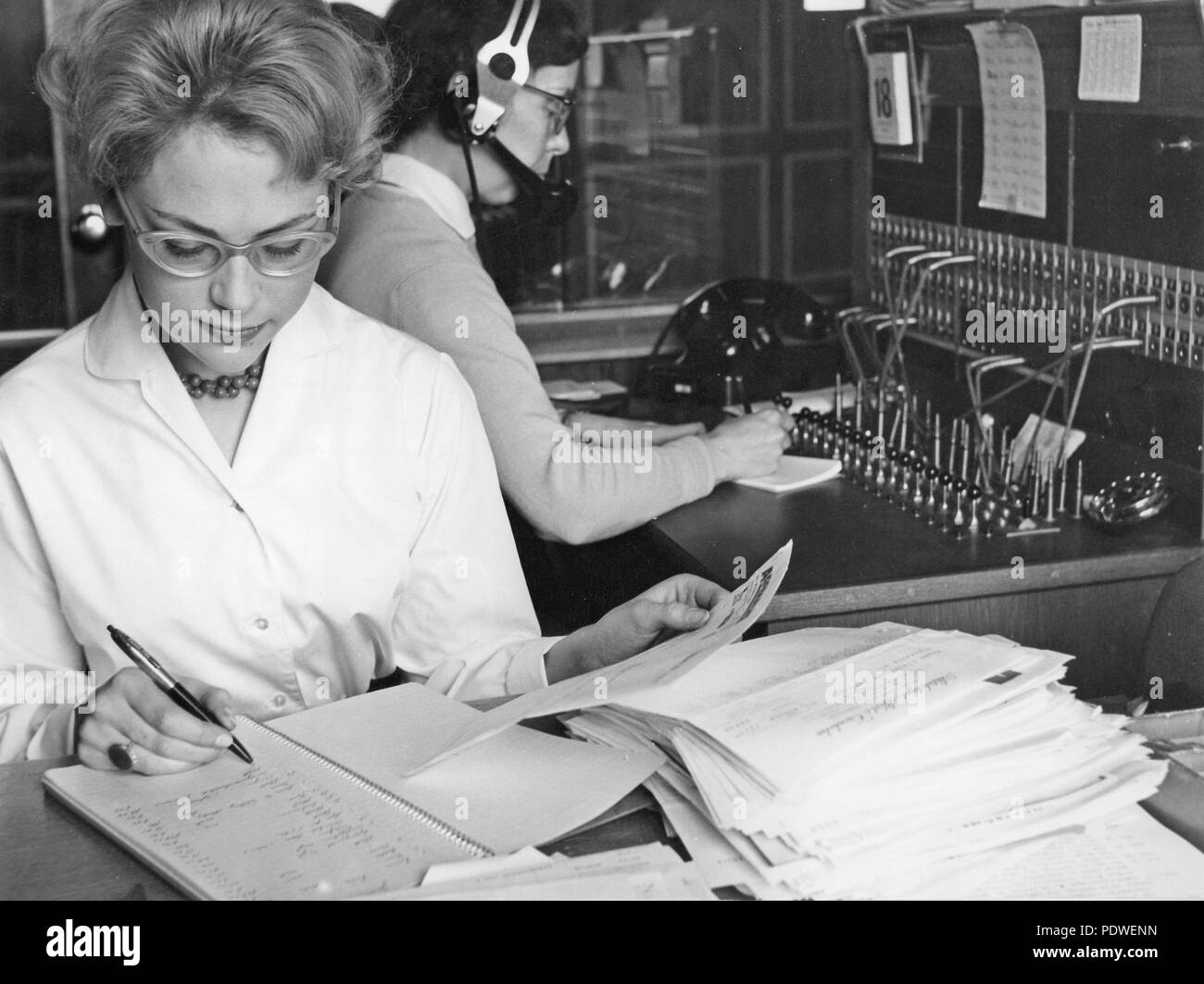 1950 Bürokauffrau und Telefon Telefonzentrale. Ein junges Büro sitzt an ihrem Schreibtisch, Bearbeitung von Dokumenten. Ein Telefon Bediener sitzt hinter das Nehmen Sie eingehende Anrufe und Weiter zu verbinden. 1958 Stockfoto