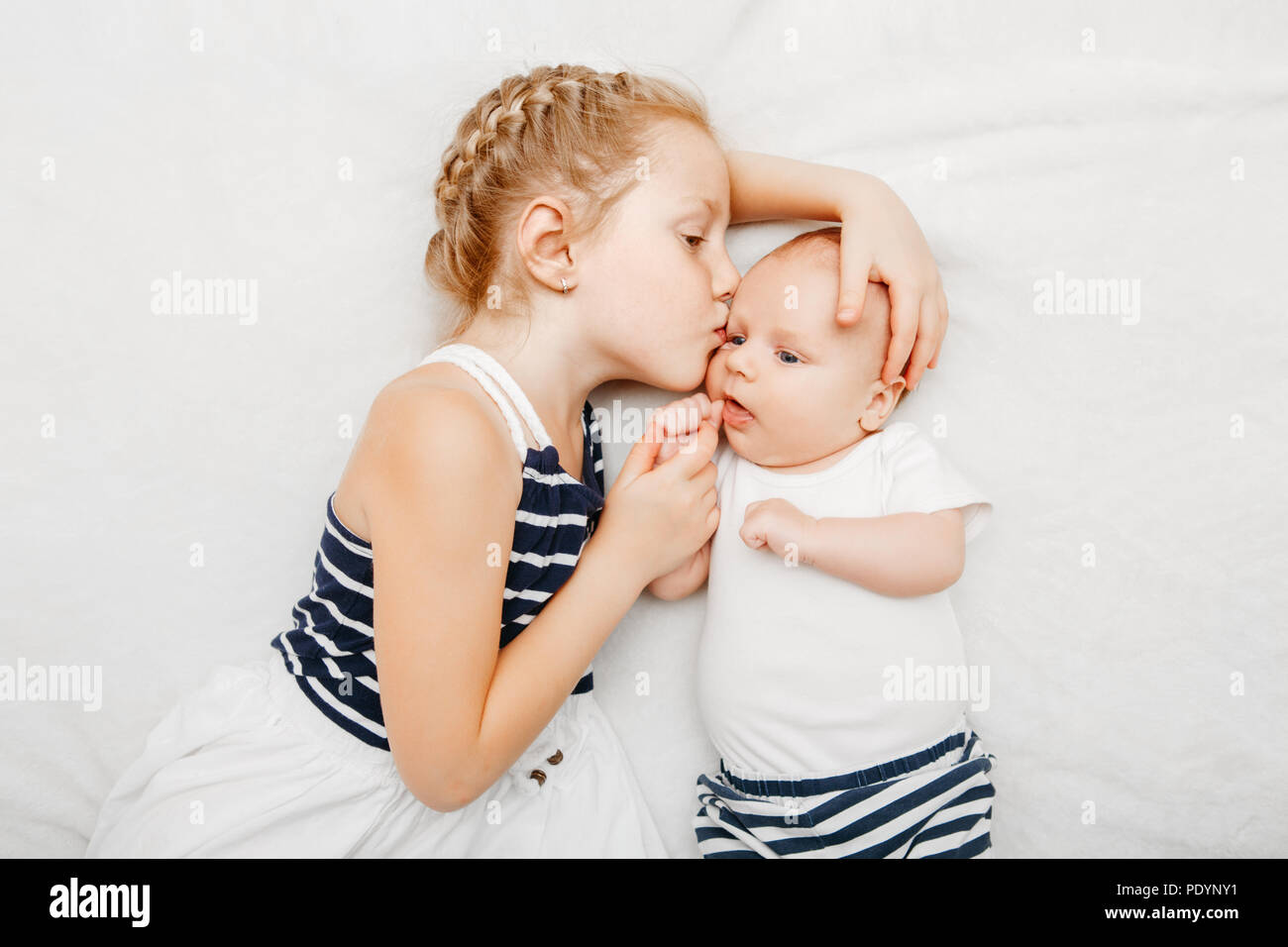 Lifestyle Portrait von niedlichen weißen Kaukasische Mädchen Schwester Holding küssen kleine Baby, liegend auf dem Bett drinnen. Ältere Geschwister mit jüngeren Bruder Neugeborenen. Stockfoto