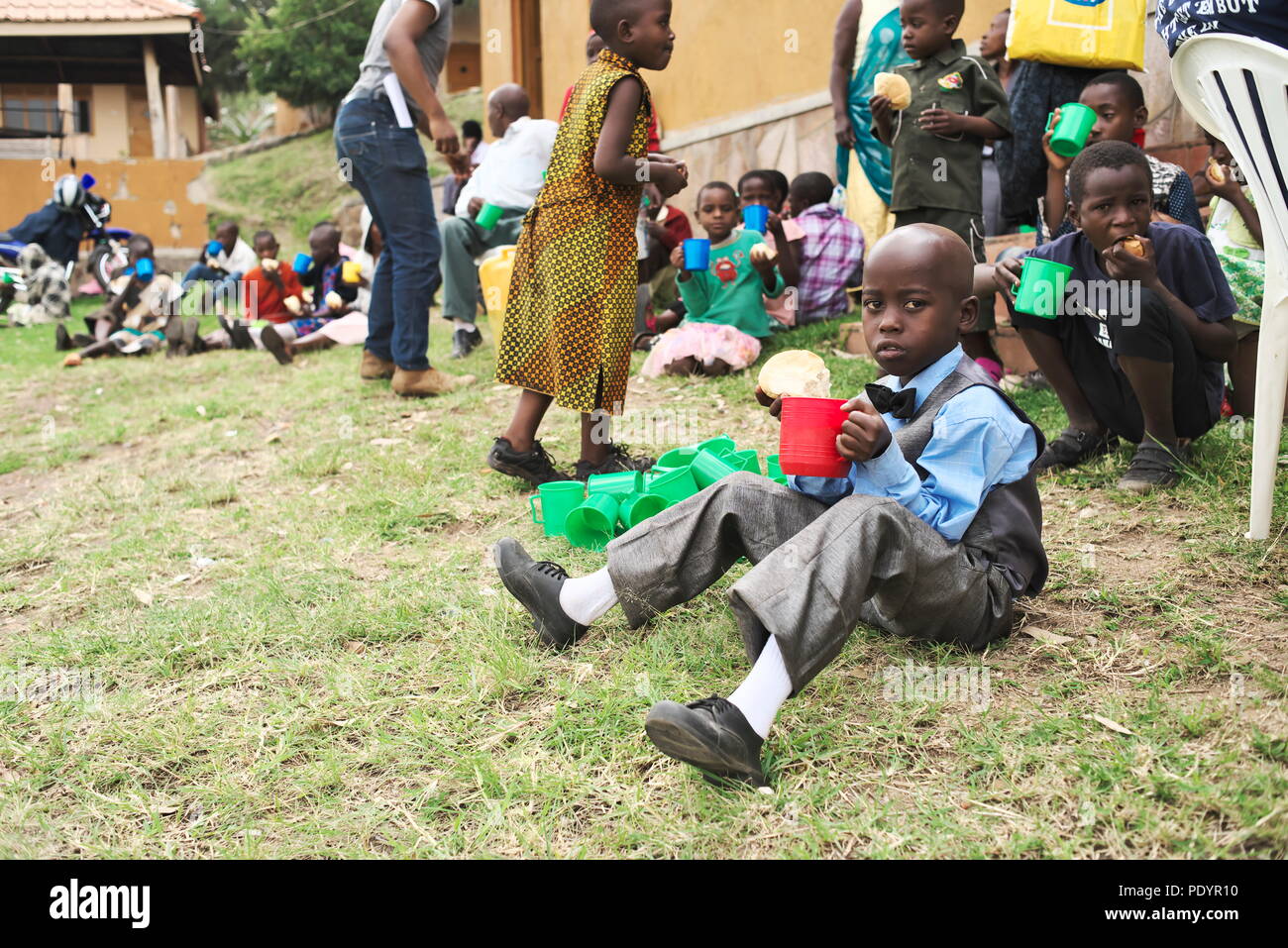Ein junges, intelligentes afrikanisches Kind sitzt auf dem Gras außerhalb eines afrikanischen Krankenhauses und isst und trinkt und trägt einen sehr eleganten Anzug Stockfoto