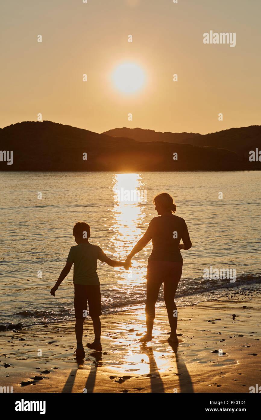 Mutter und Sohn gehen auf einem goldenen Sandstrand bei Sonnenuntergang die Sonne am Himmel und im Wasser spiegeln mit einem goldenen Glanz Stockfoto