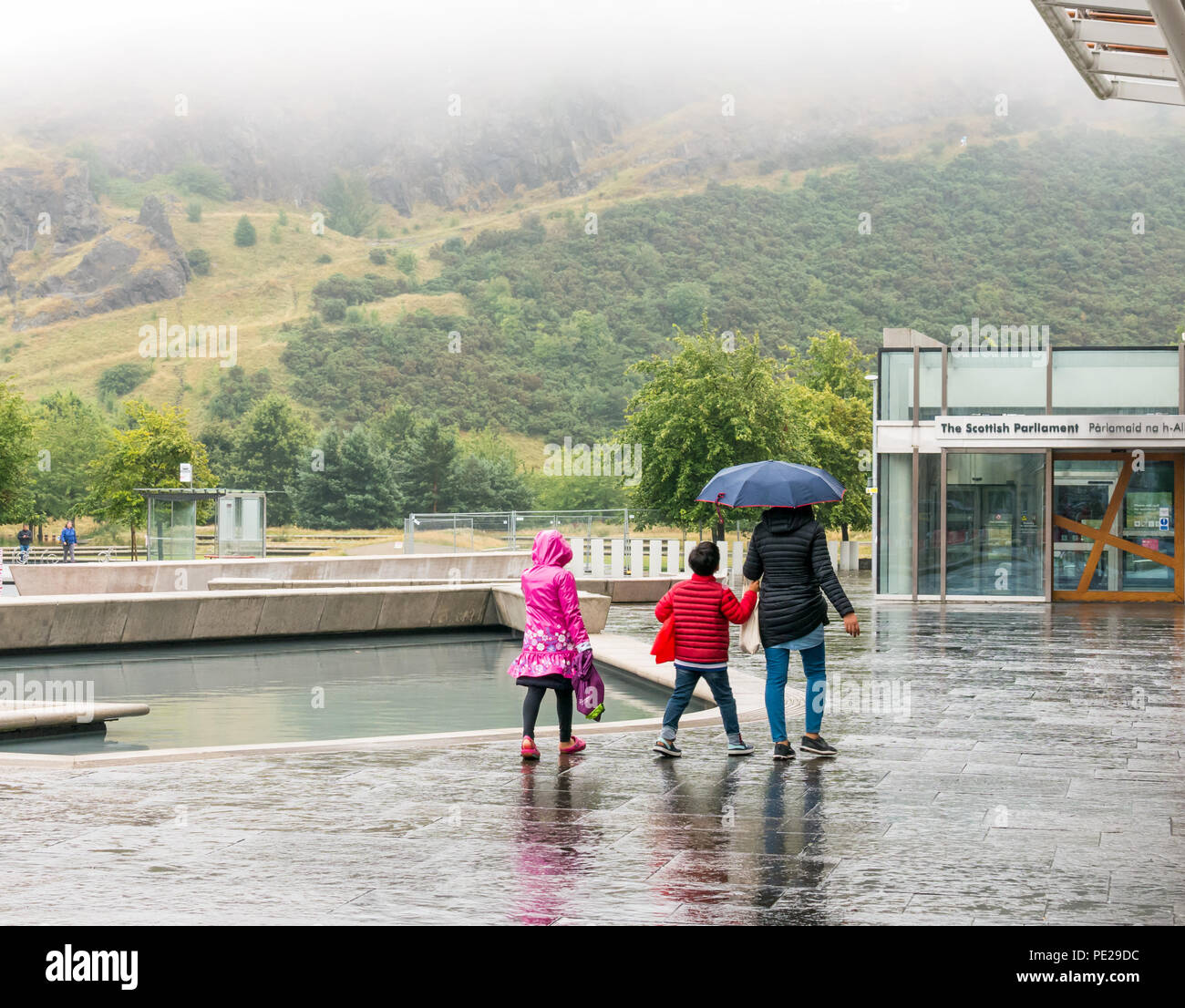 Holyrood Edinburgh, Edinburgh, Schottland, Vereinigtes Königreich, 12. August 2018. UK Wetter: Regen und Nebel, wie die Reste der Sturm Debby Hits das Kapital nicht Touristen in das schottische Parlament Gebäude mit feuchtigkeitsbeschlags auf Holyrood Park abhalten. Eltern und Kinder mit einem Regenschirm in einem hellen Regenmäntel gekleidet Stockfoto