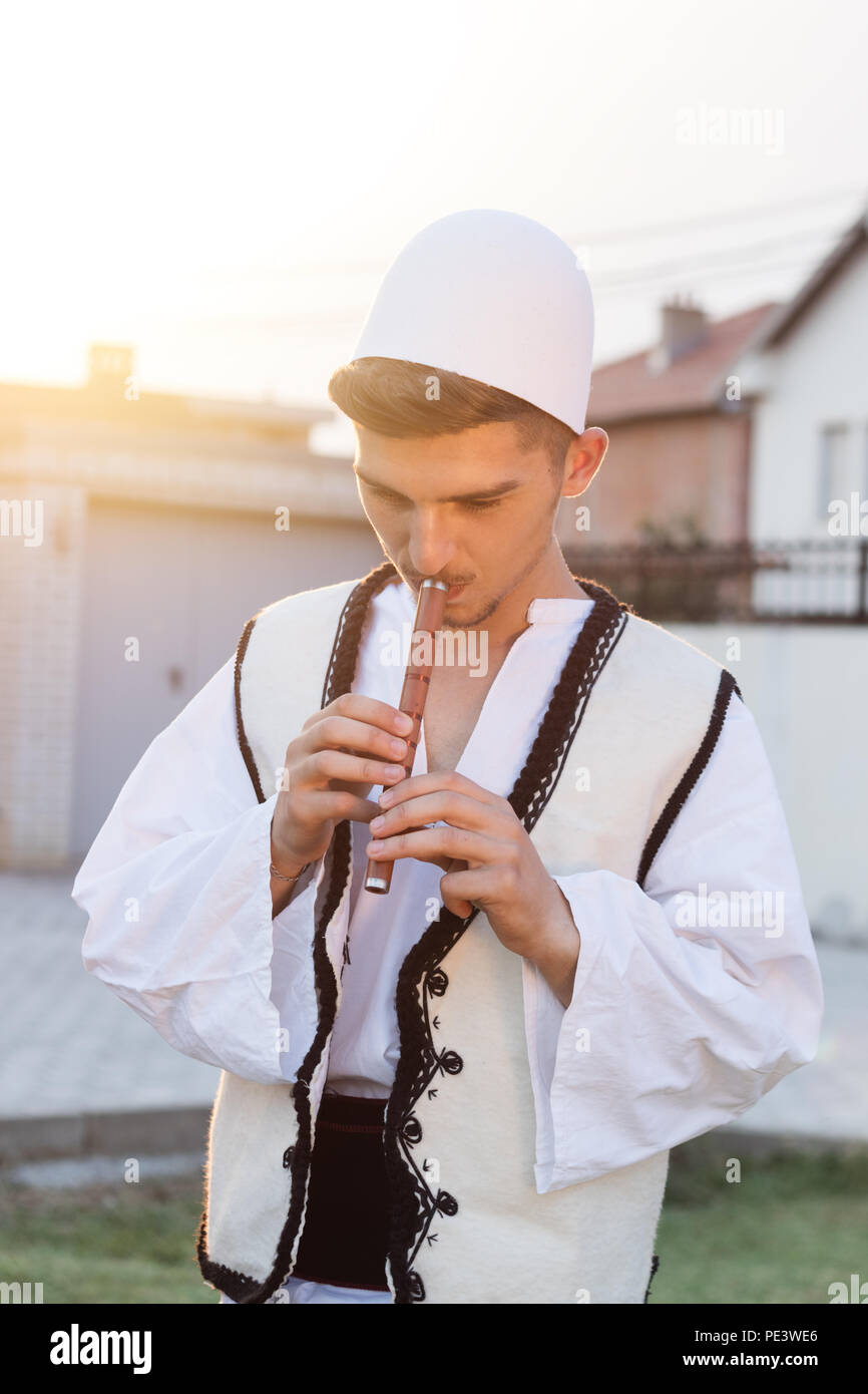 Junge Mann in der traditionellen albanischen Kostüm spielt Flöte vor der Natur Wiese und Abenddämmerung Stockfoto