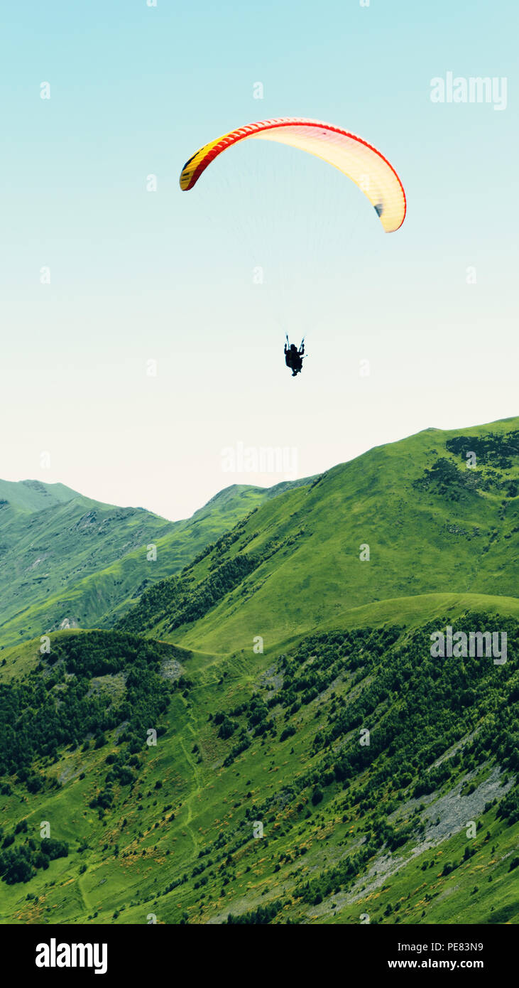 Ein Gleitschirm mit Menschen fliegt über die Berggipfel in einem strahlend blauen Himmel vor dem Hintergrund eines Berges Schlucht. Grüner Vegetation bedeckt die Mo Stockfoto