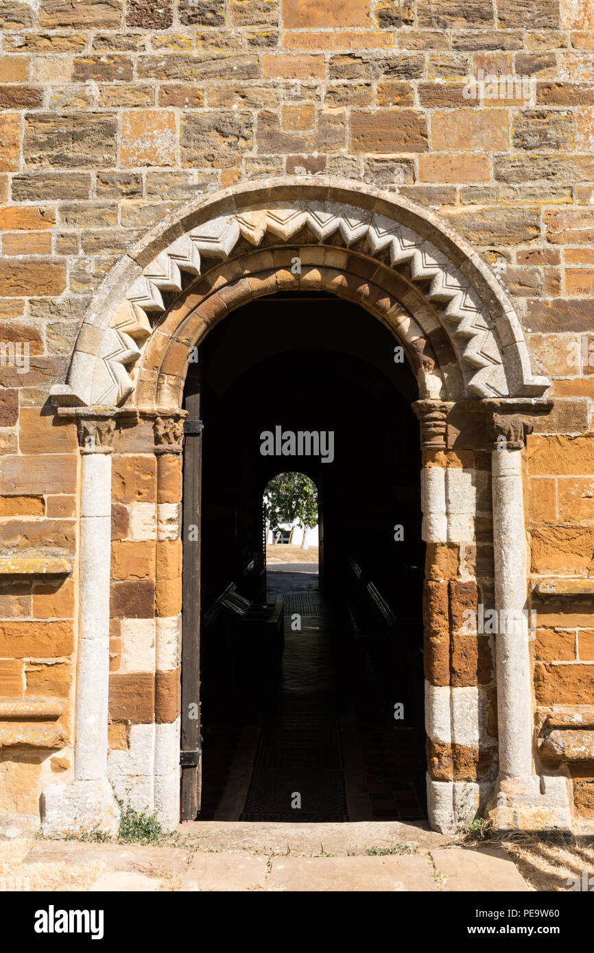Spratton, Northamptonshire, 24-07-18. Eine offene Tür in einer Wand wird von einem geschnitzten Norman arch überwunden. Im Inneren ist die Dunkelheit und eine Tür die Vermietung in Licht. Stockfoto
